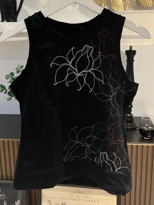 Svart linne med blommor  - Svart linne med breda axelband och blommor på framsidan i färgerna vit, grå och röd. Linnet har en sydd kant nertill och är i stretchigt material. Axelbredd: 27 cm. Längd: 54 cm. Byst: 40 cm.