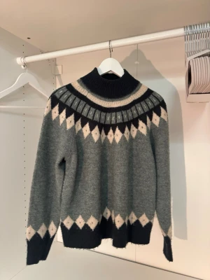 Grön stickad tröja från ONLY Studio - Snygg frönstickad tröja från ONLY Studio med mörkgrönt och vitt/beiget mönster runt halsen och nederkanten. Tröjan har hög krage, långa ärmar och ribbstickade muddar. Perfekt för kalla dagar och ger en trendig look.