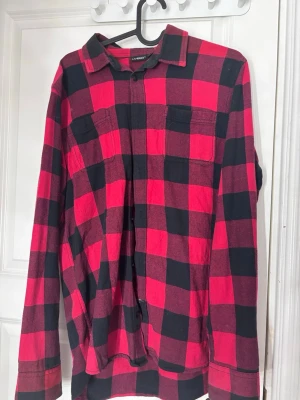 Röd och svart rutig skjorta - Klassisk röd och svart rutig skjorta från Livergy med långa ärmar och en bröstfickor. Skjortan har knappar framtill och en avslappnad passform, perfekt för en cool och avslappnad stil. Finns ett hål o nån fläck på höger arm 