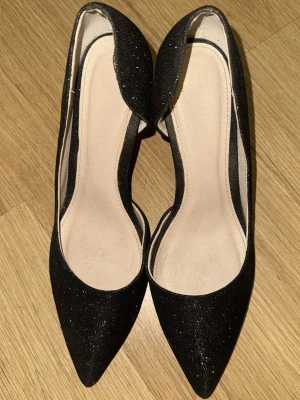 Svarta glittriga pumps från Tiamo - Snygga svarta pumps från Tiamo med glittrigt tyg och spetsig tå. Skorna har låg klack och öppen sida för en trendig look. Perfekta för dig som vill ha något extra till outfiten.