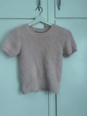 Ljusrosa stickad t-shirt från Zara - Mysig ljusrosa stickad t-shirt från Zara med korta ärmar och rund halsringning. Tröjan har en fluffig, mjuk struktur och är perfekt för lager på lager eller som ett statement-plagg. Passar dig som gillar en söt och trendig stil.
