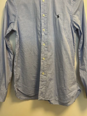 Ljusblå skjorta från Ralph Lauren - Klassisk ljusblå skjorta från Ralph Lauren med knappstängning framtill och broderad logga på bröstet. Skjortan har lång ärm och är tillverkad i bomull, perfekt för en clean och stilren look. Knappt använd 