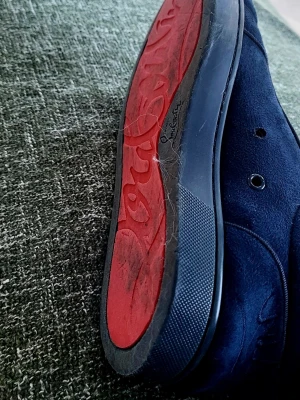 Mörkblå sneakers från Christian Louboutin - Snygga mörkblå sneakers från Christian Louboutin med den ikoniska röda sulan. Skorna har en klassisk rund tå och är tillverkade i mocka med svarta detaljer. Perfekta för dig som vill sticka ut med exklusiv stil.