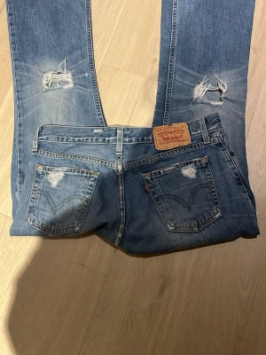 Levi's 517 ljusblå bootcut jeans - Säljer ett par ljusblå Levi's 517 jeans med bootcut passform. Jeansen har slitna detaljer och hål på både fram- och baksida, samt klassiska fem fickor och Levi's patch bak. Perfekta för en avslappnad och trendig look.W.30 L.34