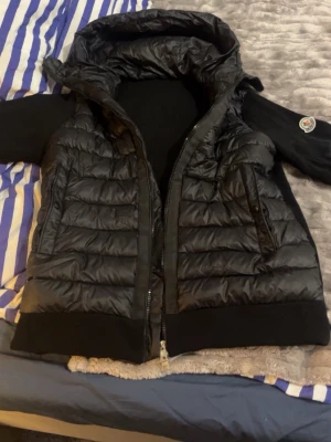 Svart Cardigan från Moncler med huva - Snygg svart Cardigan från Moncler med stickade partier och vadderade sektioner. Västen har en stor huva, dragkedja framtill och praktiska fickor med dragkedja. Moncler-logga på Armen, ger en exklusiv känsla. Perfekt för lager-på-lager och streetwear.