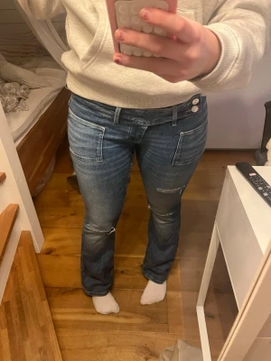 Blå bootcut jeans med fickor fram - Säljer ett par blå bootcut jeans med två stora fickor fram och dubbla knappar i midjan. Jeansen har slitna detaljer på benen och en låg midja. Perfekta för en avslappnad och trendig look.