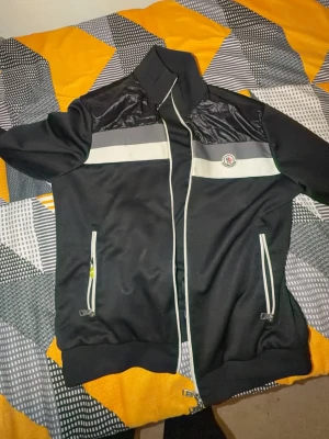 Svart vindjacka från Moncler XL - Snygg svart vindjacka från Moncler med vita detaljer och grått blockmönster över bröstet. Jackan har hög krage, dragkedja framtill och två fickor med vita dragkedjor. Moncler-logga på bröstet och ribbade muddar vid ärmar och midja. ÄKTA