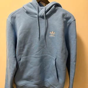 Ljusblå hoodie från Adidas - Snygg ljusblå hoodie från Adidas med klassisk huva och dragsnören. Framtill finns en stor magficka och det ikoniska Adidas-loggan i vitt på bröstet. Perfekt för en avslappnad och sportig stil.