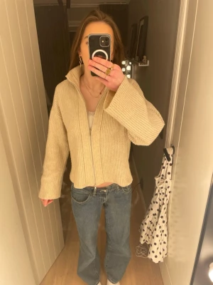 Beige stickad ziptröja med krage - Beige stickad tröja med dragkedja och hög krage. Tröjan har raka, vida ärmar och är ribbstickad i ett mjukt material. Perfekt för lager-på-lager och enkel att styla till jeans eller kjol.