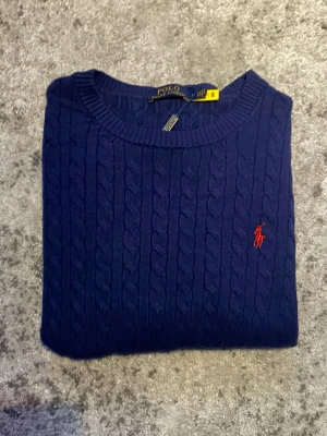 Mörkblå Ralph Lauren tröja  - Säljer denna klassiska kabelstickade Ralph Lauren tröjan i mörkblå med röd logga. Storleken är L fast passar mer som en mindre M enligt mig, tröjan är dessutom i mycket fint skick. Hör gärna av dig vid frågor om t.ex. mått, bilder etc!