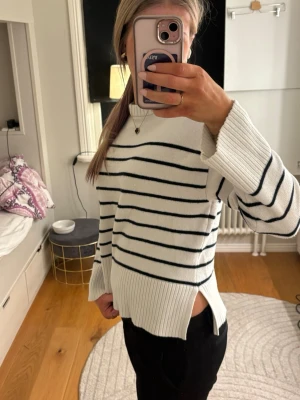 Vit tröja med svarta ränder - Vit tröja med svarta horisontella ränder och breda ribbade muddar. Tröjan har slitsar i sidorna och en avslappnad passform. Perfekt för en stilren och klassisk look. Är i storlek S och har inga defekter