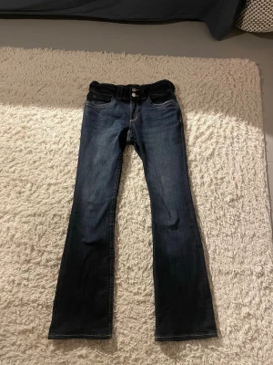 Mörkblå bootcut jeans med knappar - Snygga mörkblå jeans med bootcut-modell och dubbla knappar i midjan. Jeansen har klassiska fickor fram och bak, samt dekorativa knappar på bakfickorna. Perfekta för en trendig och avslappnad look.
