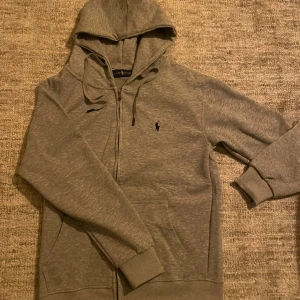 Grå hoodie från Ralph Lauren - Klassisk grå hoodie från Ralph Lauren med dragkedja framtill och justerbar huva med snörning. Ikoniska lilla Polo-loggan broderad på bröstet. Tillverkad i mjuk bomullsmix och har ribbade muddar vid ärmar och nederkant. Perfekt för en chill och stilren look.