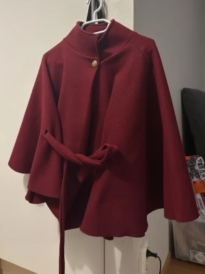Vinröd kappa med bälte och knapp - Stilren vinröd kappa med hög krage, dekorativ guldknapp och knytbälte i midjan. Kappan har en vid, svepande passform och är perfekt för höst och vinter. Enkel men elegant design som lyfter varje outfit.