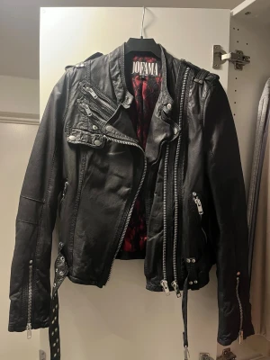 Svart skinnjacka från Jofama - Cool svart skinnjacka från Jofama med flera dragkedjor, bälte i midjan och detaljerade fickor. Jackan har en klassisk bikerstil och röd innerfoder. Perfekt för dig som vill ha en edgy look.