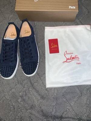 Mörkblå sneakers från Louboutin - Snygga mörkblå sneakers från Christian Louboutin med rund tå och vita platta sulor. Skorna har snörning och är dekorerade med svarta nitar framtill för en edgy look. Tillverkade i mocka och levereras med originalpåse och box.