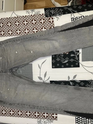 Grå bootcut/flare jeans som är custom made. - Storlek: W29 L34 Snygga grå jeans med slitningar och små hål på benen. Jeansen har en klassisk femficksmodell och är tillverkade i denim. Perfekta för en avslappnad och trendig look.