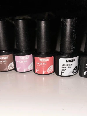 MTSSII gelnagellack & nagelstickers - Flera flaskor MTSSII Color Gel nagellack i olika färger (brun, rosa, röd, svart) samt två ark nagelstickers – ett med guldiga symboler och ett med färgglada motiv som hjärtan, stjärnor och nallar. Perfekt för att skapa unika nageldesigner hemma.