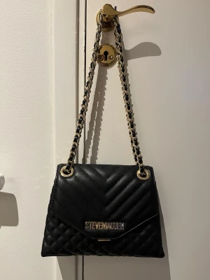 Svart quiltad handväska Steve Madden - Stilren svart quiltad handväska från Steve Madden med kedjerem i guld och svart. Väskan har ett elegant mönster och metallogo framtill. Perfekt accessoar för att lyfta din outfit. 