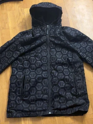 Svart vindjacka med mönster från Philipp Plein - Snygg svart vindjacka från Philipp Plein med heltryckt logomönster och huva. Jackan har dragkedja framtill och två fickor med dragkedja. Perfekt för dig som vill sticka ut med en exklusiv streetstil.