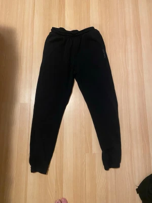 Ett par svarta byxor från Pilago  - Svarta mjukisbyxor från Pilago med elastisk midja och fickor på sidorna. Byxorna har en enkel och clean design med diskret logga på ena benet. Perfekta för chill dagar eller när du vill vara bekväm men ändå se snygg ut.