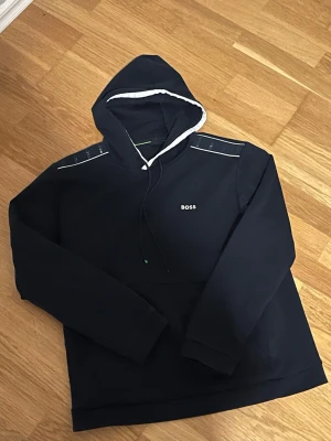 BOSS Hoodie - Snygg mörkblå hoodie från BOSS med vit logga på bröstet och vita detaljer längs huvan och axlarna. Klassisk känguruficka framtill och justerbar dragsko i huvan. Perfekt för en avslappnad och trendig stil. Storleken är L men passar M bättre