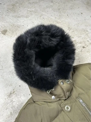 Olivgrön dunjacka med svart fuskpäls - Snygg olivgrön dunjacka från Moose Knuckles med svart fuskpäls runt huvan. Jackan har quiltad design, dragkedjor och knappar framtill samt fickor med lock och ärmficka. Perfekt för kalla vinterdagar och har en modern, oversized passform.