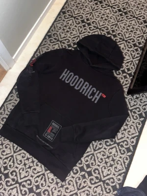 Svart HOODRICH hoodie med tryck - Svart hoodie från Hoodrich med stor logga på bröstet och detaljerade tryck på fickan och ryggen. Klassisk huva och känguruficka framtill. Snygga röda och vita detaljer som ger en streetwear vibe.