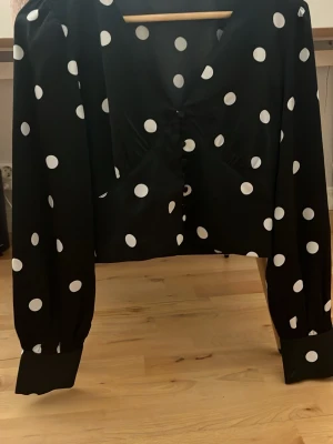 Svart prickig blus med knappar - Trendig svart blus med stora vita prickar och snygga knappar framtill. Blusen har lång ärm med breda muddar och en lätt v-ringad krage. Perfekt för dig som gillar retrostil och vill sticka ut lite extra.