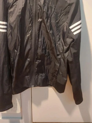 Svart vindjacka från Adidas Respons - Snygg svart vindjacka från Adidas med klassiska vita ränder på ärmarna och dragkedja framtill. Lätt och smidig modell i polyester, perfekt för blåsiga dagar. Adidas-logga på bröstet och Response-kollektionen.