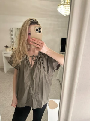 Olivgrön blus med knytning i halsen - Snygg olivgrön blus med v-ringning och knytning vid halsen. Blusen har korta, vida ärmar. 