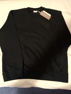 Svart sweatshirt från dior - Snygg svart sweatshirt från dior med diskret logga broderad på bröstet. Klassisk rund halsringning och ribbade muddar vid ärmslut och nederkant. Tillverkad i mjukt material för en bekväm känsla. Perfekt till jeans eller joggers.