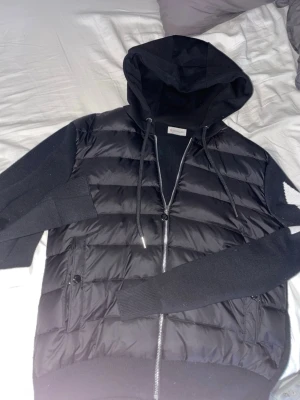 Äkta Svart Moncler pufferjacka med huva - Reupload (pga att jag vill ha den såld) PRISET KAN DISKUTERAS VID SNABB AFFÄR. Säljer den pga att jag ej gillar den längre! Hej, jag säljer min Svarta pufferjacka (cardigan) från Moncler med stickade ärmar och huva. Jackan har dragkedja framtill, snörning vid huvan och coolt zigzag-mönster i svart och vitt på ena ärmen. Fickor med knappstängning och Moncler-logga på ärmen. Perfekt för dig som vill ha en snygg och varm jacka. Storleken är M men passar också S då den är lite liten vid midjan !