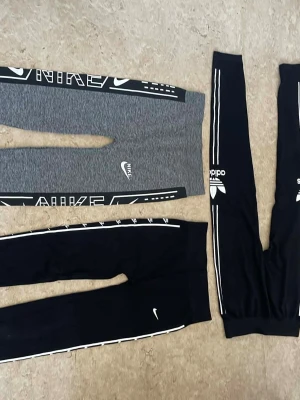 Nike och Adidas leggings med logga - Tre par leggings, två från Nike och ett från Adidas. Ett par är gråmelerade med svartvita detaljer och stor Nike-logga, de andra två är svarta med vita ränder och loggor längs benen. Alla har tight passform och sportig look, perfekta för träning eller chill.