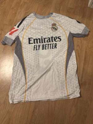 Real Madrid Mbappé fotbollströja - Säljer en Real Madrid fotbollströja med Mbappé och nummer 9 på ryggen. Tröjan är vit med grå och gula detaljer, mönstrad med diskreta hexagoner och har svarta tryck. Ränder på ärmslut och flera sponsorer, bland annat Emirates och HP. Perfekt för fotbollsfans!