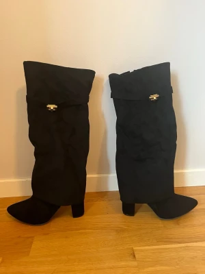 Svarta stövlar med gulddetalj - Säljer ett par svarta stövlar med bred skaft och snygg guldfärgad spänndetalj upptill. De har klack och är gjorda i mocka för en lyxig känsla. Perfekta att styla med kjol eller jeans för en cool look.