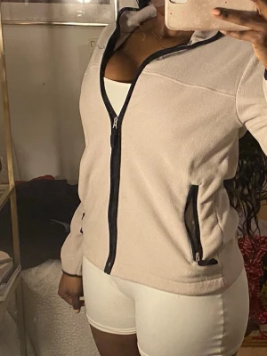 Beige fleecejacka med svarta detaljer - Mysig beige fleecejacka med svarta dragkedjor och detaljer. Jackan har hög krage och två fickor med dragkedja framtill. Perfekt för lager-på-lager och chill dagar. Enkel och stilren design som passar till det mesta.