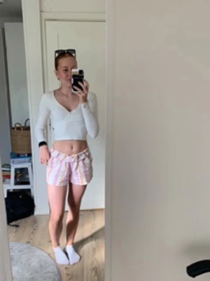 Randiga shorts från Vero Moda  - Säljer ett par luftiga shorts från Vero Moda i storlek XS. De är vita med breda ränder i ljusrosa och beige, har hög midja och bälteshällor. Materialet är linneblandning, perfekt för varma sommardagar.