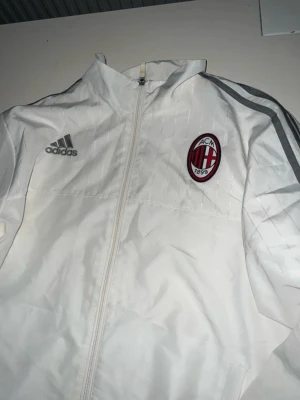 Vit Adidas AC Milan vindjacka - Säljer en vit vindjacka från Adidas med AC Milan-logga på bröstet och klassiska grå ränder längs ärmarna. Jackan har hel dragkedja, ståkrage och en röd detalj längst ner. Perfekt för dig som gillar fotboll och vill ha en sportig stil.