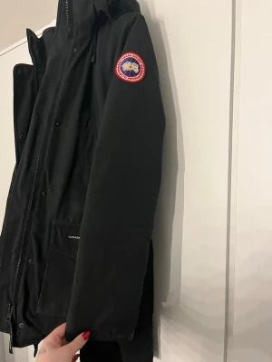 Svart dunjacka från Canada Goose - Svart dunjacka från Canada Goose med huva och klassisk logga på ärmen. Jackan har långa ärmar, dragkedja framtill och stora fickor med lock. Perfekt för kalla dagar och har en stilren look som funkar till allt. Päls finns också, tyvärr har jackan blivit för liten för mig!