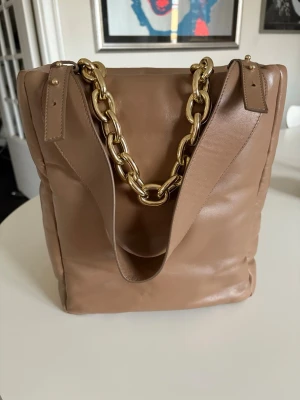 Beige axelväska från Stand Studio - Säljer en beige axelväska från Stand Studio i mjukt skinn med chunky guldfärgad kedja som handtag. Väskan har en stilren, fyrkantig form och ett innerfack med dragkedja. Perfekt för dig som gillar minimalistisk design och snygga detaljer. Väskan har två olika handtag.