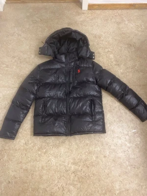 Svart dunjacka från Polo Ralph Lauren - Svart dunjacka från Polo Ralph Lauren med röd broderad logga på bröstet. Jackan har huva, två sidofickor med dragkedja och en quiltad design. Materialet är glansigt och känns riktigt chill för kalla dagar. Perfekt för dig som vill ha en snygg och varm jacka.