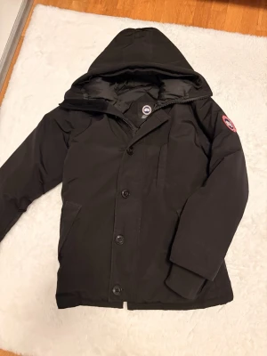 Canada goose chateau parka  - Svart dunjacka från Canada Goose med huva och klassisk logga på ärmen. Jackan har knappar och dragkedja framtill, två fickor och ribbade muddar vid ärmslut. Insidan är quiltad och jackan är perfekt för kalla dagar. Snygg och stilren design som håller dig varm.