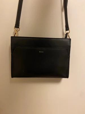 Svart axelväska från Wera i skinn - Snygg svart axelväska från Wera i slätt skinn med guldfärgade detaljer. Väskan har ett stilrent, rektangulärt format och flera fack inuti, inklusive dragkedja. Perfekt storlek för det viktigaste och enkel att matcha med olika outfits. Använd 3 gånger och nypris är 800kr