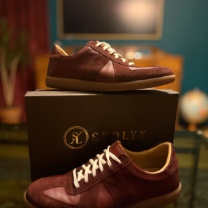 Vinröda sneakers från Skolyx - Säljer ett par stilrena vinröda sneakers från Skolyx med snygga kontraster mellan mocka och skinn. Skorna har vita snören, rund tå och beige innersula. Yttersulan är gummifärgad och har ett klassiskt mönster. Perfekt för dig som gillar en clean look med lite edge.