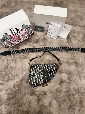 Dior Saddle handväska med bred axelrem - Säljer en ikonisk Dior Saddle handväska i svart och beige med klassiskt Dior-mönster. Väskan har en bred, broderad axelrem med metall-detaljer och Christian Dior Paris-tryck. Materialet är bomull och skinn, med snygga guldfärgade metalldetaljer och D-spänne framtill. Pris can diskuteras