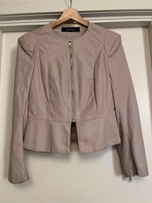Zara Beige peplum skinnjacka - Beige peplum skinnjacka från Zara med dragkedjor vid ärmsluten och framtill. Jackan har en kortare, figurnära passform och är i skinnimitation. JÄTTESNYGG! Använd väldigt sparsamt då jag älskade modellen och ville spara på den, välomhändertagen men har defekt vid kragen då skinnimitationen spruckit (med tiden tyvärr). 