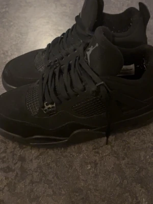 Svarta Nike Jordan 4 sneakers - Säljer ett par svarta Nike Jordan 4 sneakers med snygga meshpaneler och klassisk Jumpman-logga på plösen och hälen. Skorna har snörning, platt sula och är tillverkade i mocka och syntet. Perfekt för dig som gillar stilrena och ikoniska sneakers.