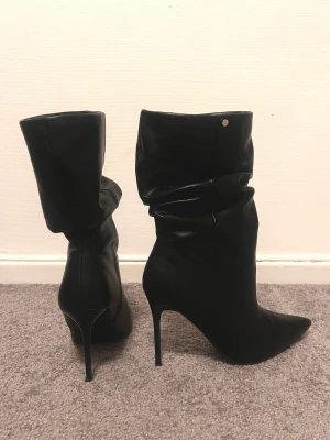Svarta stövletter med spetsig tå - Säljer ett par svarta stövletter med hög klack och spetsig tå. Skorna har en snygg slouchy design och är tillverkade i skinnimitation. Perfekta för att lyfta din outfit med en edgy vibe.