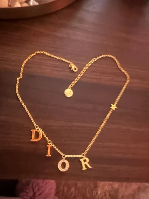 Guldfärgad Dior halsband med bokstäver och silver halsband dior  - Säljer ett snyggt halsband från Dior i guldfärg med kedja och hängande bokstäver som stavar DIOR. Bokstaven O är dekorerad med små glittrande stenar och det finns även en liten stjärndetalj på kedjan. Perfekt accessoar för att lyfta din outfit.